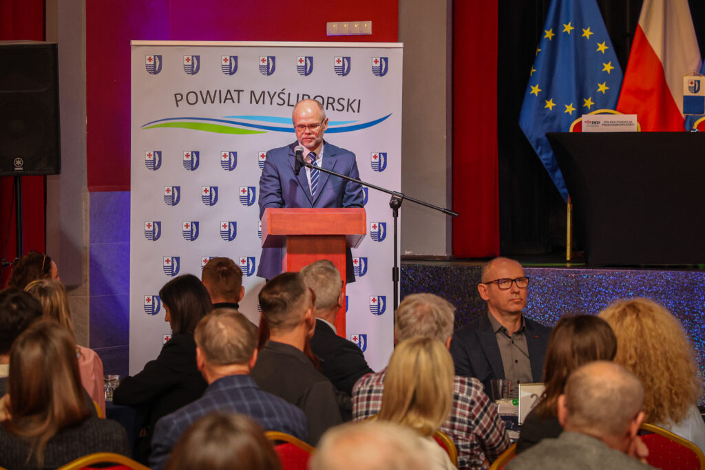 Mężczyzna przemawia przy mównicy w tle ścianka z herbami i napis Powiat Myśliborski; komunikat dotyczy wystąpienia lub wydarzenia organizowanego przez Powiat Myśliborski.