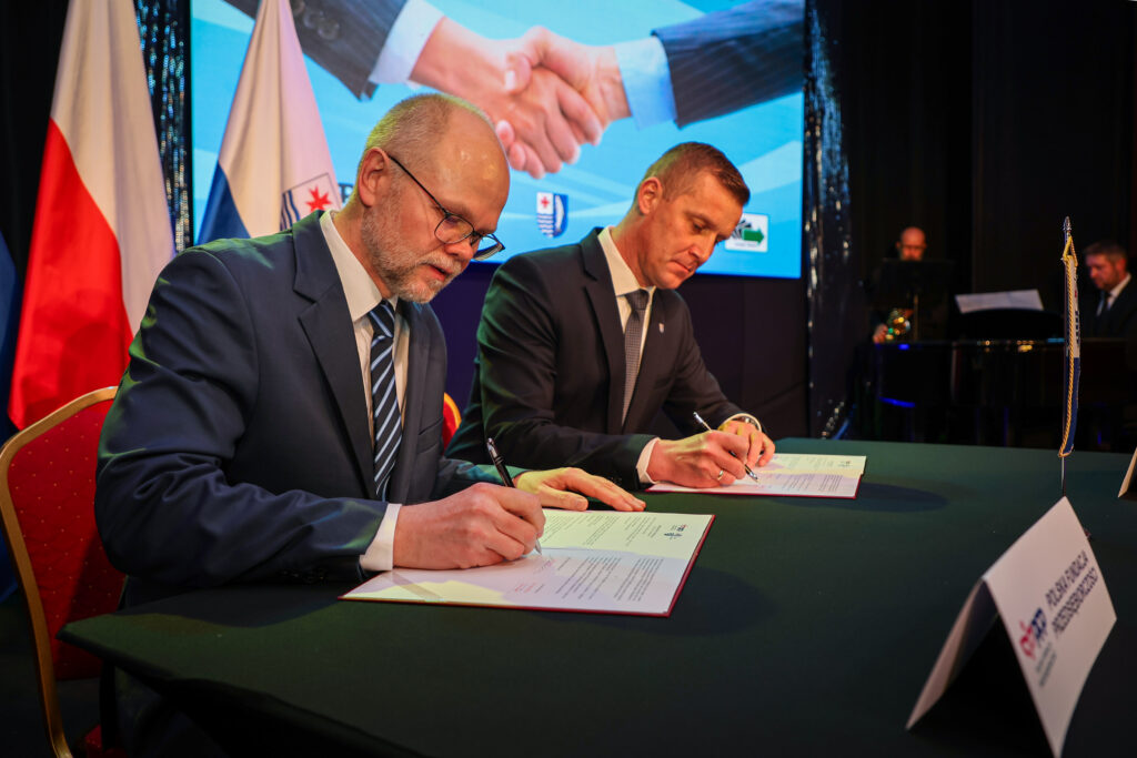 Dwóch mężczyzn w garniturach podpisuje dokumenty przy stole, w tle na ekranie widoczny uścisk dłoni oraz stoją flagi Polski i samorządu. Podpisanie umowy o współpracy między instytucjami.