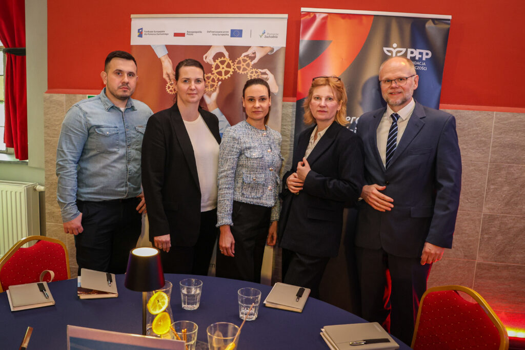 Pięć osób w strojach biznesowych stoi przy stole konferencyjnym na tle dwóch roll-upów i komunikuje spotkanie lub współpracę w obszarze funduszy i przedsiębiorczości, na lewym banerze napis Fundusze Europejskie dla Pomorza Zachodniego i grafika zębatek w dłoniach, na prawym logo PFP Polska Fundacja Przedsiębiorczości