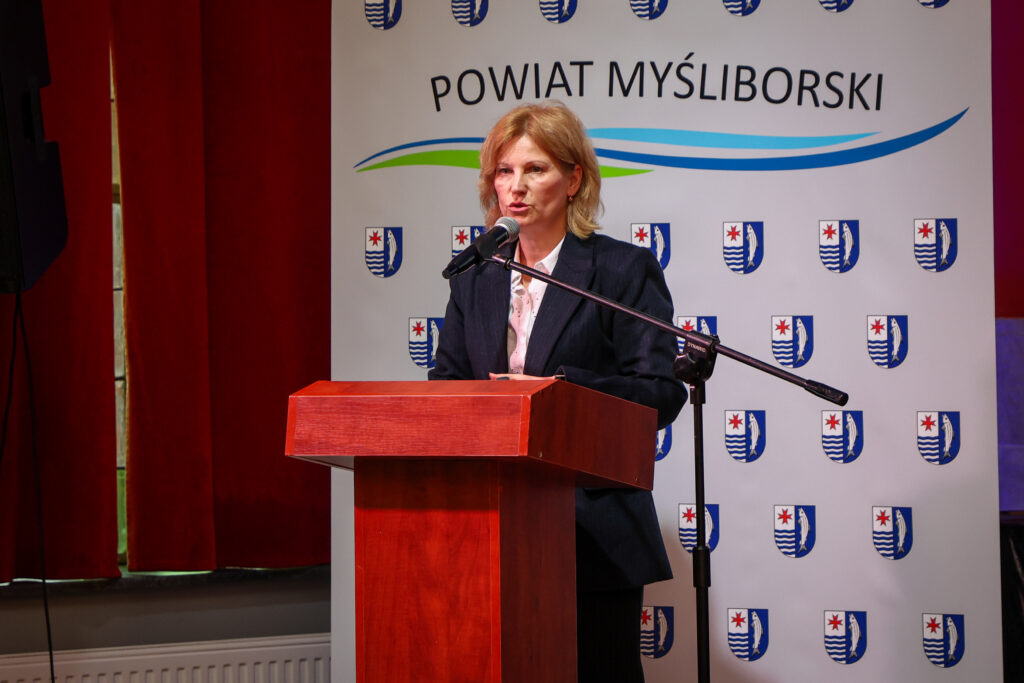 Kobieta przemawia przy mównicy, w tle ścianka z herbami i napis Powiat Myśliborski; komunikat dotyczy wystąpienia lub wydarzenia organizowanego przez Powiat Myśliborski.