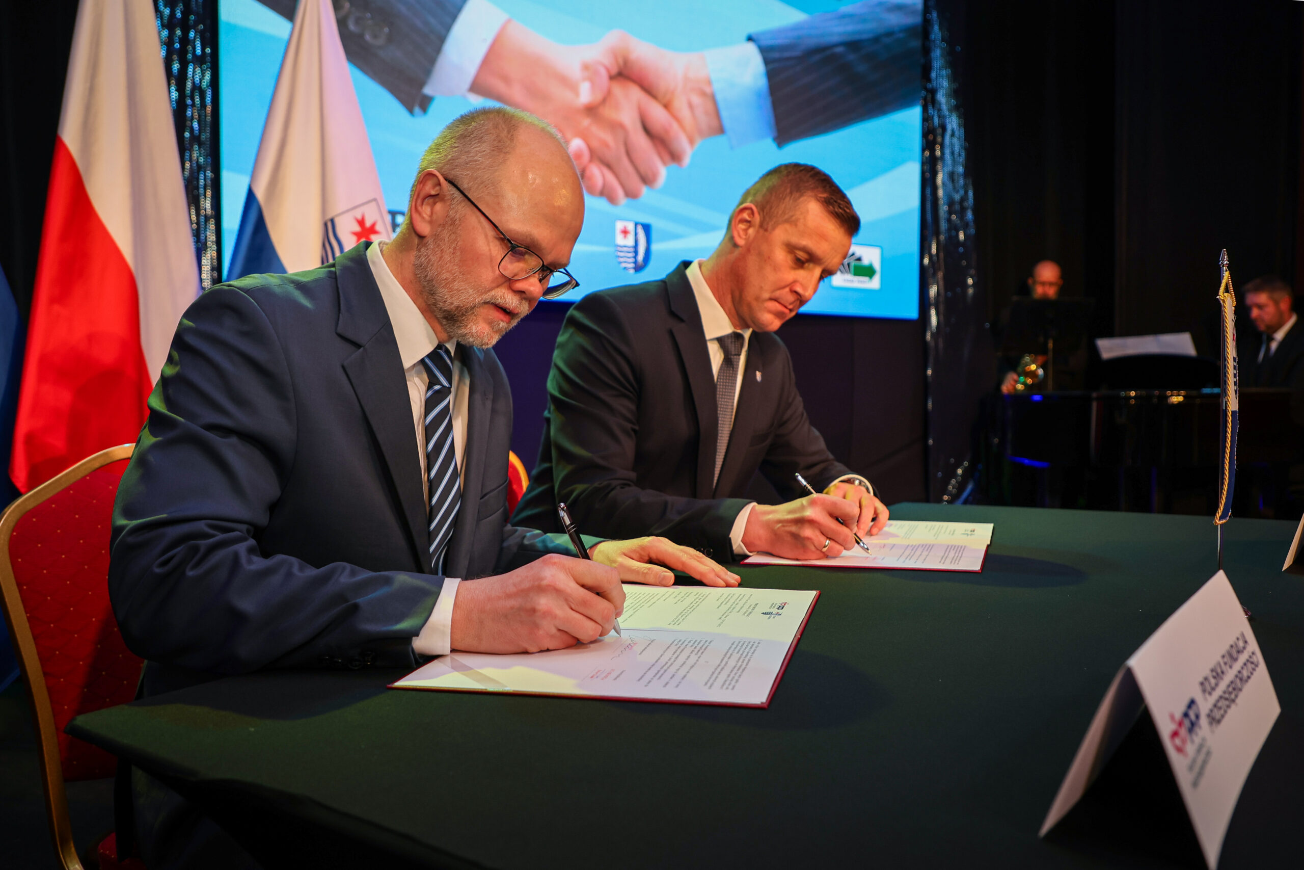 Dwóch mężczyzn w garniturach podpisuje dokumenty przy stole, w tle na ekranie widoczny uścisk dłoni oraz stoją flagi Polski i samorządu. Podpisanie umowy o współpracy między instytucjami.