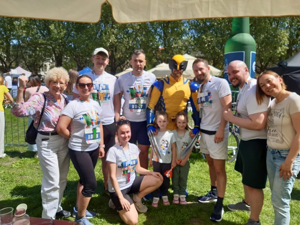 Grupa uczestników Biegu Sztafet Firmowych pozuje do wspólnego zdjęcia na trawie, w centrum stoi osoba przebrana za Wolverine’a, obok dwoje dzieci i uczestnicy z medalami oraz numerami startowymi. W tle widoczne namioty i strefa wydarzenia plenerowego.