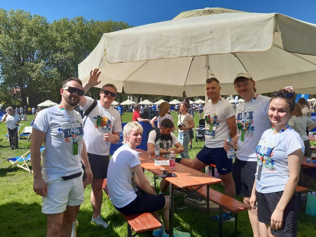 Grupa uczestników Biegu Sztafet Firmowych stoi i siedzi przy drewnianym stole pod dużym parasolem, część osób pokazuje medale i numery startowe, w tle trwa plenerowe wydarzenie sportowe. Na koszulkach widoczny napis Bieg Sztafet Firmowych 4 × 4 km.