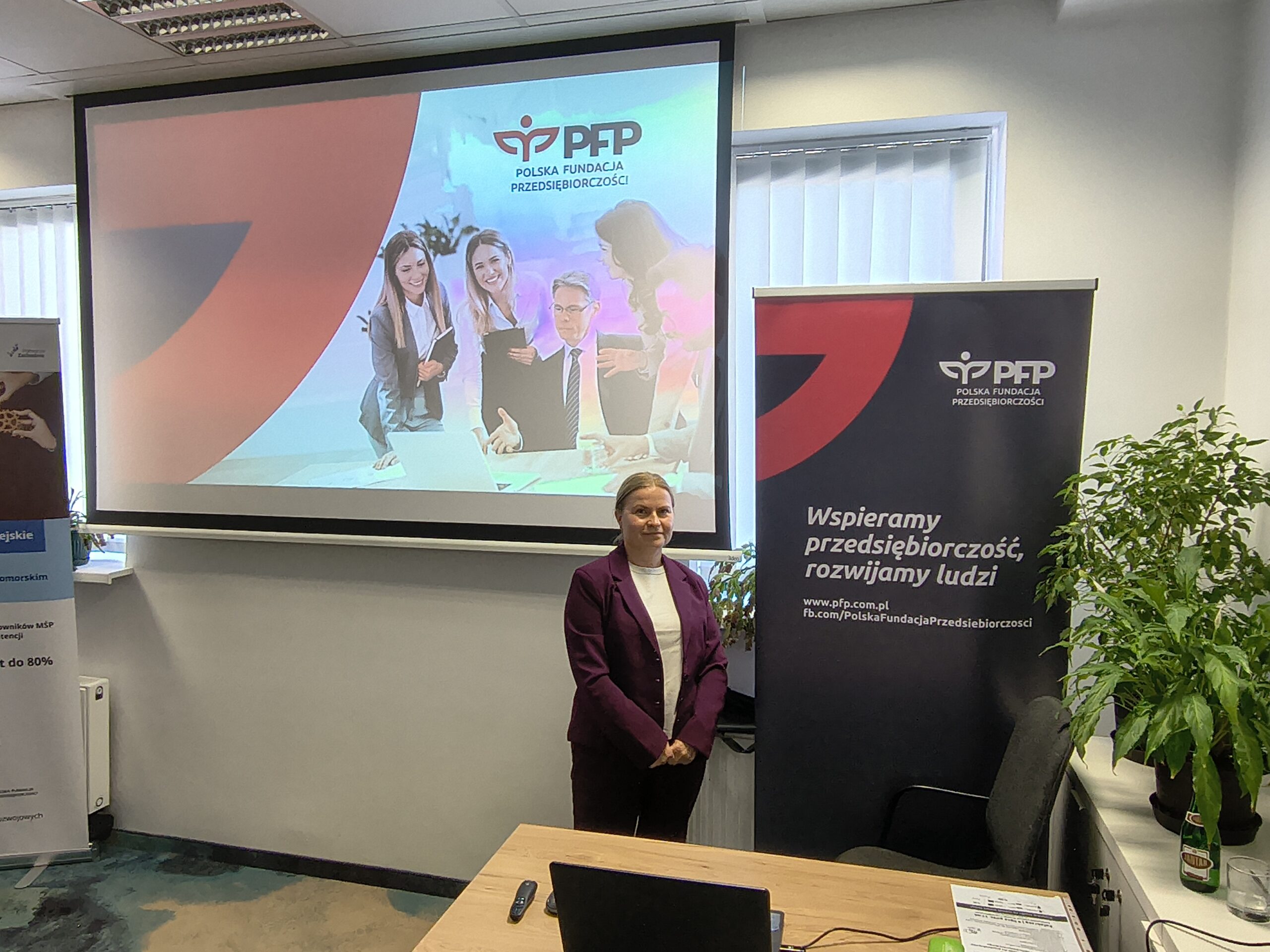 1000010744 Kobieta stoi przed ekranem z prezentacją i roll-upem Polskiej Fundacji Przedsiębiorczości. Trwa spotkanie informacyjne, na ekranie widoczne jest logo PFP Polska Fundacja Przedsiębiorczości, a na roll-upie hasło: Wspieramy przedsiębiorczość, rozwijamy ludzi.