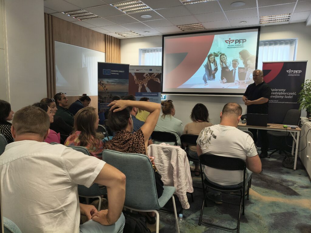 Uczestnicy siedzą na sali konferencyjnej i słuchają prezentacji prowadzonej podczas spotkania informacyjnego. Na ekranie wyświetlono logo PFP Polska Fundacja Przedsiębiorczości, obok znajduje się roll-up z hasłem: Wspieramy przedsiębiorczość, rozwijamy ludzi.