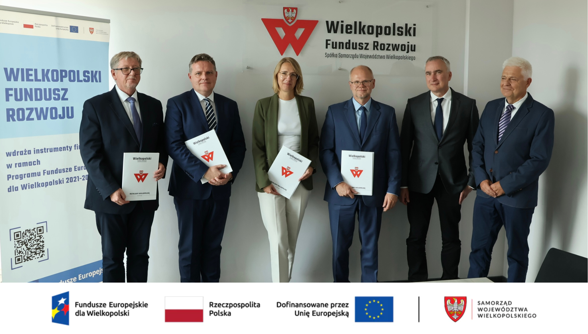 Sześć osób stoi w siedzibie Wielkopolskiego Funduszu Rozwoju, część trzyma firmowe teczki; baner informuje o wdrażaniu instrumentów finansowych w ramach Programu Fundusze Europejskie dla Wielkopolski 2021–2027. Na samym dole od lewej strony widnieją loga: Fundusze Europejskie dla Wielkopolski, Barwy Rzeczypospolitej Polskiej, logo Unii Europejskiej z dopiskiem Dofinansowane przez Unię Europejską, pionowa czarna linia oddzielająca, Samorząd Województwa Wielkopolskiego.